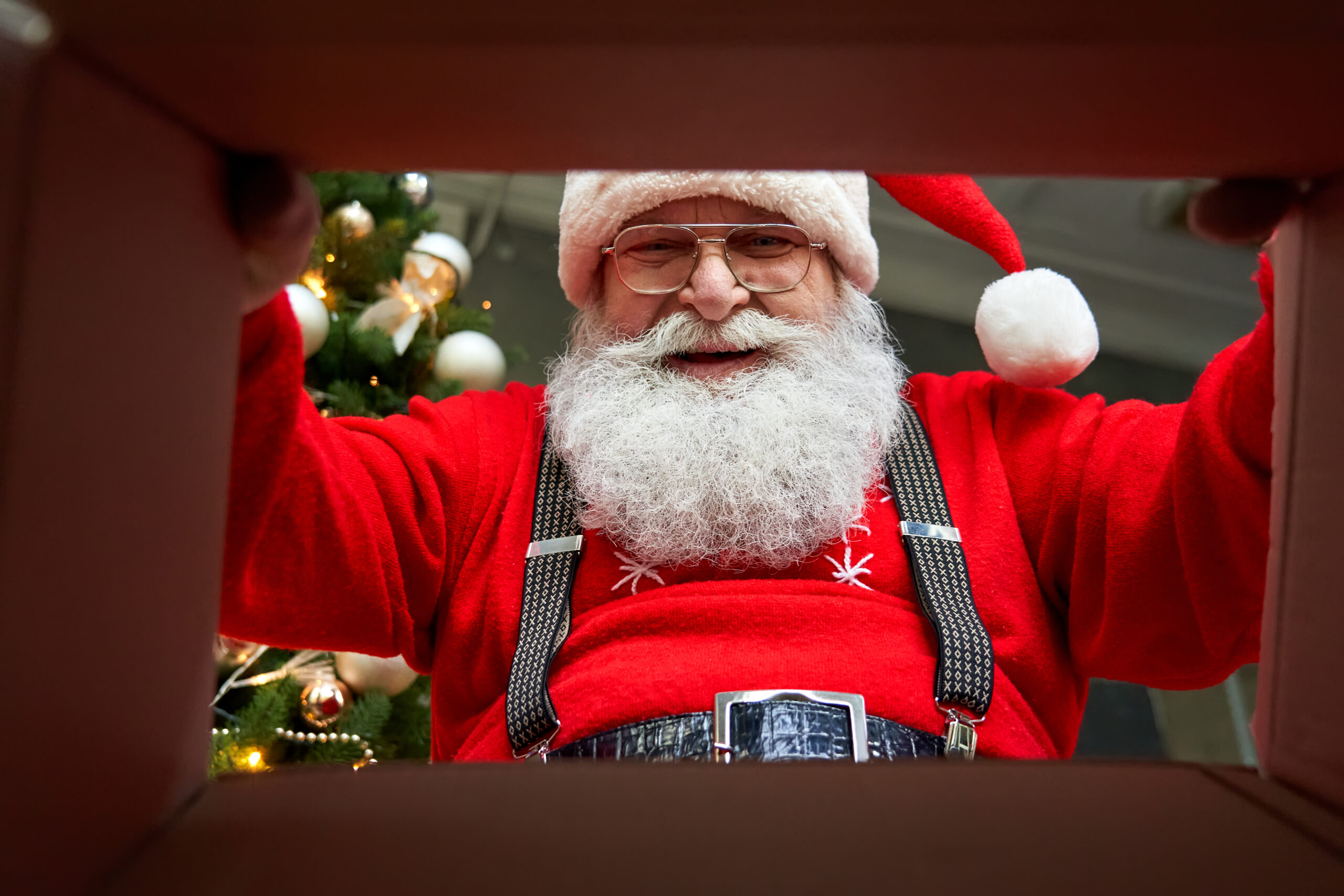 Happy old bearded Santa Claus wearing hat packing present looking inside cardboard box wrapping gift preparing package delivery on xmas eve. Merry Christmas surprise concept, close up view from below.